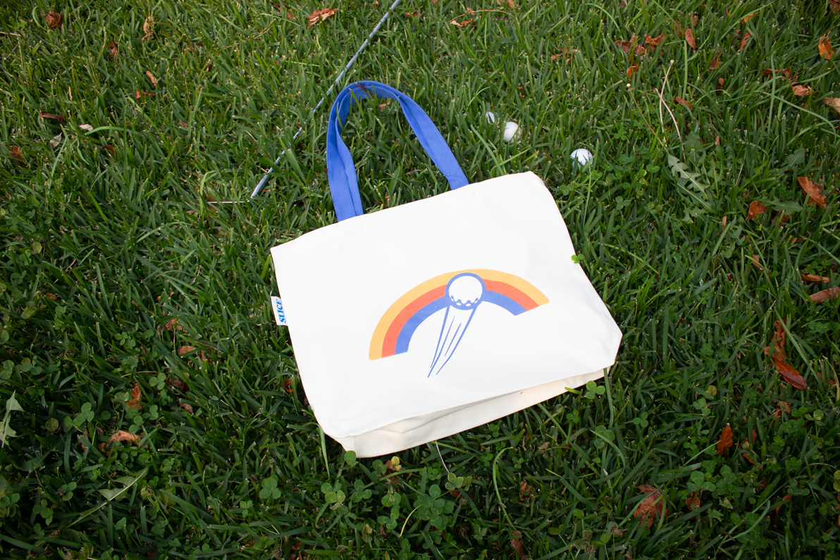 Organic cotton tote back in off-white with Slice Golf rainbow logo and blue handle on grass.