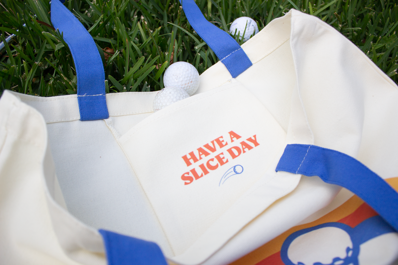 Organic cotton tote back in off-white with Slice Golf rainbow logo and blue handle on grass.